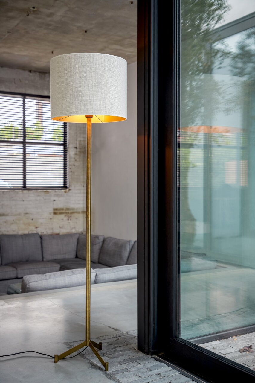 Stehlampe mit weißem zylindrischem Lampenschirm und goldenem Ständer, in einem modernen Wohnzimmer mit Blick auf ein Fenster.