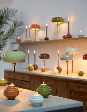 Tischleuchte SURIN in verschiedenen Farben auf einem Tisch, mit einer grünen Lampe im Vordergrund und mehreren Lampen im Hintergrund, seitliche Perspektive.