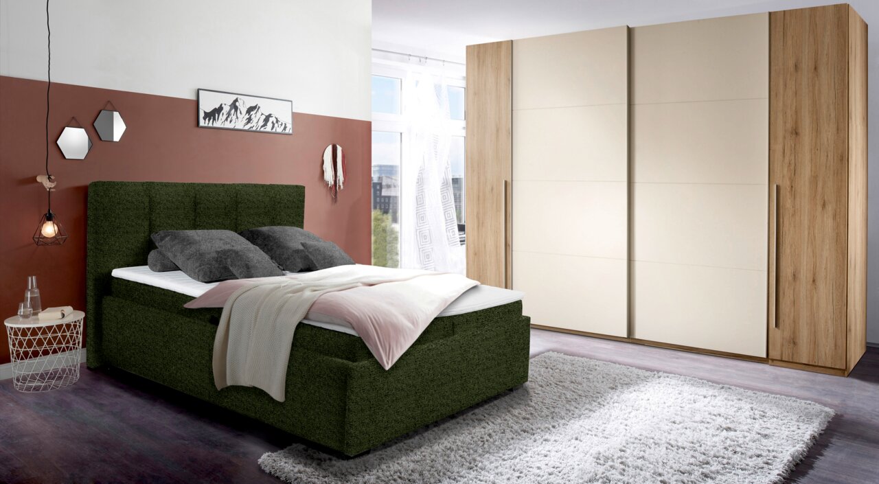 Polsterbett in einem modernen Schlafzimmer, seitliche Perspektive, mit grünem Stoffbezug, dekoriert mit Kissen und Decken, neben einem großen Kleiderschrank.