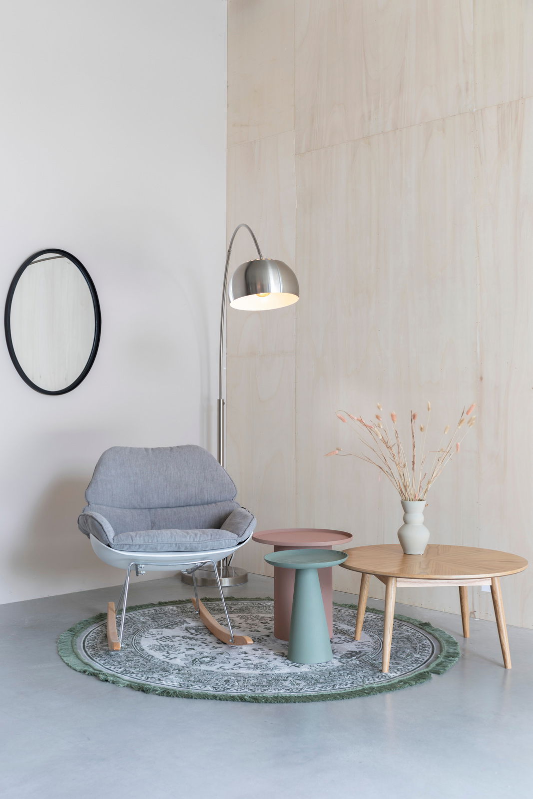 Ein stilvoll eingerichtetes Wohnzimmer mit einem grauen Schaukelstuhl, zwei modernen Beistelltischen in Rosa und Grün, einer Stehlampe aus Metall und einem runden Teppich. Perspektive: Frontalansicht.