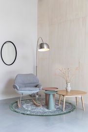 Ein stilvoll eingerichtetes Wohnzimmer mit einem grauen Schaukelstuhl, zwei modernen Beistelltischen in Rosa und Grün, einer Stehlampe aus Metall und einem runden Teppich. Perspektive: Frontalansicht.