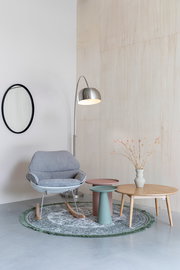 Ein stilvoll eingerichtetes Wohnzimmer mit einem grauen Schaukelstuhl, zwei modernen Beistelltischen in Rosa und Grün, einer Stehlampe aus Metall und einem runden Teppich. Perspektive: Frontalansicht.