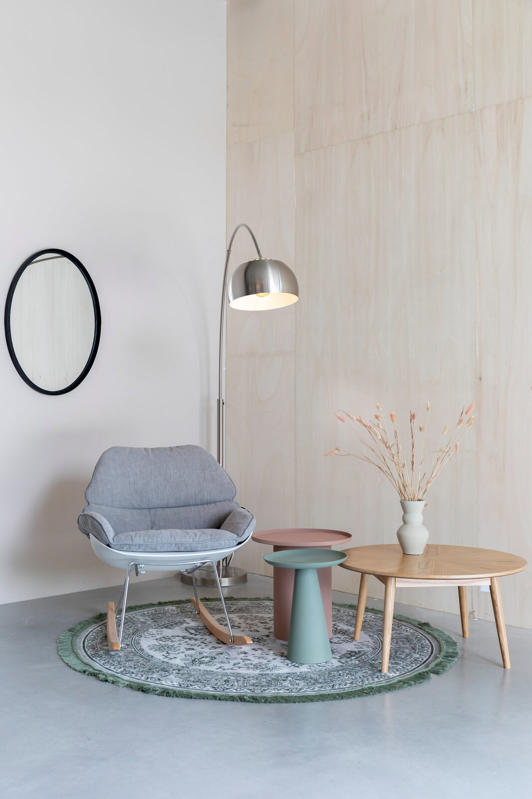 Ein stilvoll eingerichtetes Wohnzimmer mit einem grauen Schaukelstuhl, zwei modernen Beistelltischen in Rosa und Grün, einer Stehlampe aus Metall und einem runden Teppich. Perspektive: Frontalansicht.