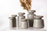 Eine Gruppe von grauen Vasen der Serie Imperia in verschiedenen Größen, auf einem weißen Tisch arrangiert. Die Vasen haben eine moderne, minimalistische Form und eine matte Oberfläche. Eine Vase ist mit getrockneten Gräsern dekoriert. Perspektive: seitlic