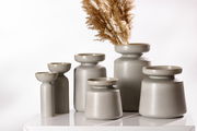 Eine Gruppe von grauen Vasen der Serie Imperia in verschiedenen Größen, auf einem weißen Tisch arrangiert. Die Vasen haben eine moderne, minimalistische Form und eine matte Oberfläche. Eine Vase ist mit getrockneten Gräsern dekoriert. Perspektive: seitlic