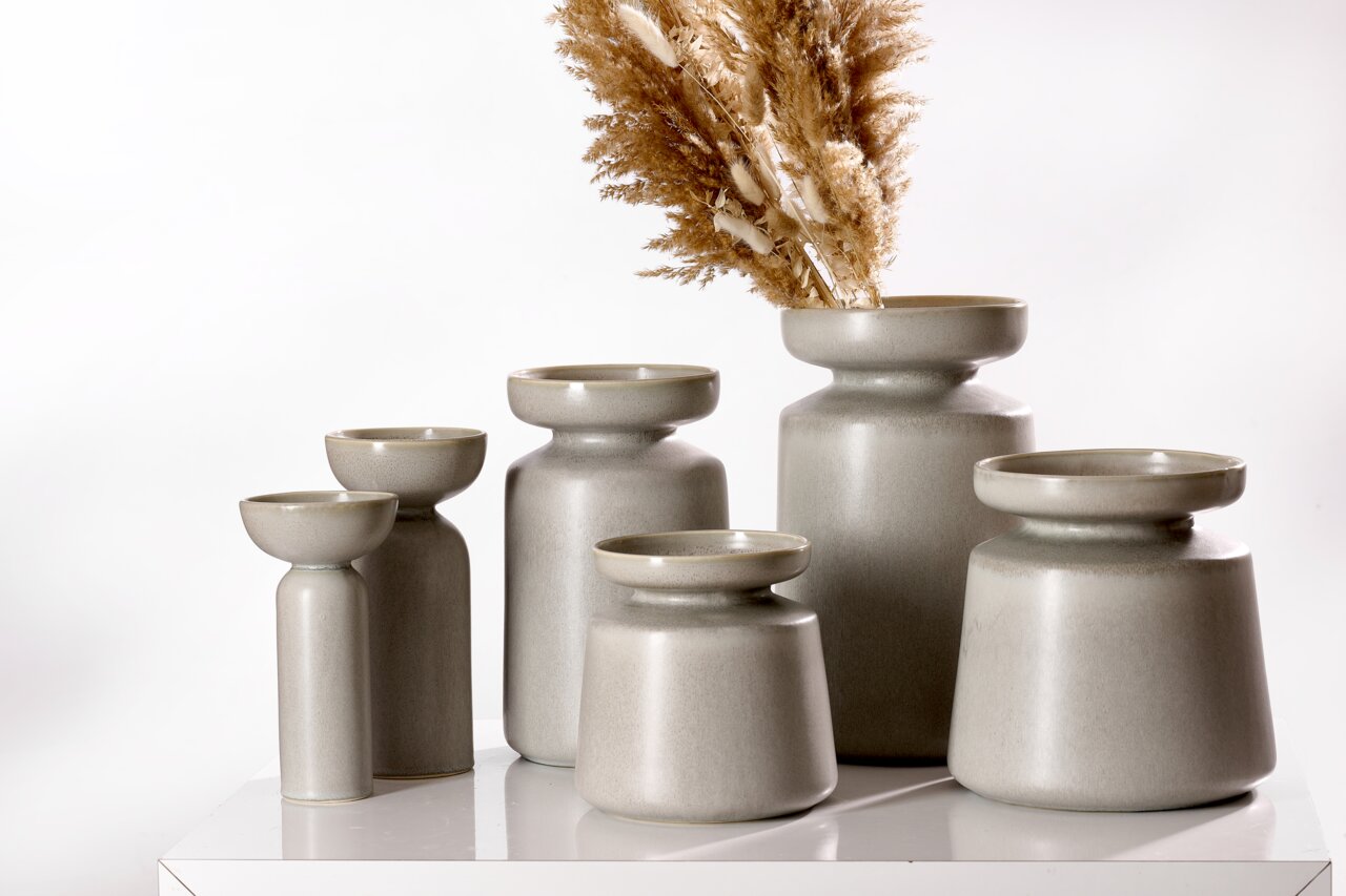 Eine Gruppe von grauen Vasen der Serie Imperia in verschiedenen Größen, auf einem weißen Tisch arrangiert. Die Vasen haben eine moderne, minimalistische Form und eine matte Oberfläche. Eine Vase ist mit getrockneten Gräsern dekoriert. Perspektive: seitlic