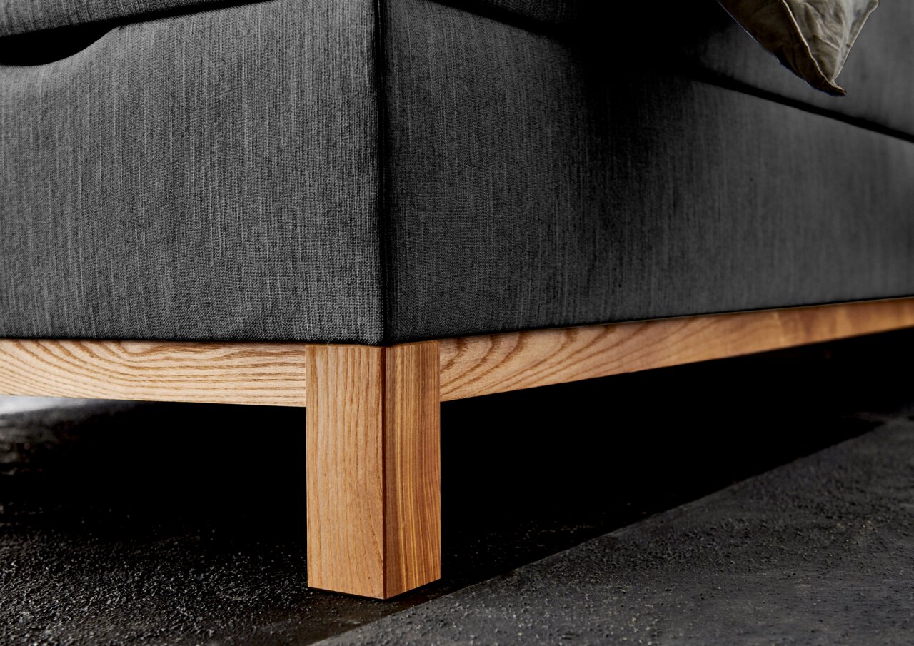 Nahaufnahme eines Boxspringbettes mit grauem Stoffbezug und Holzfüßen, seitliche Perspektive