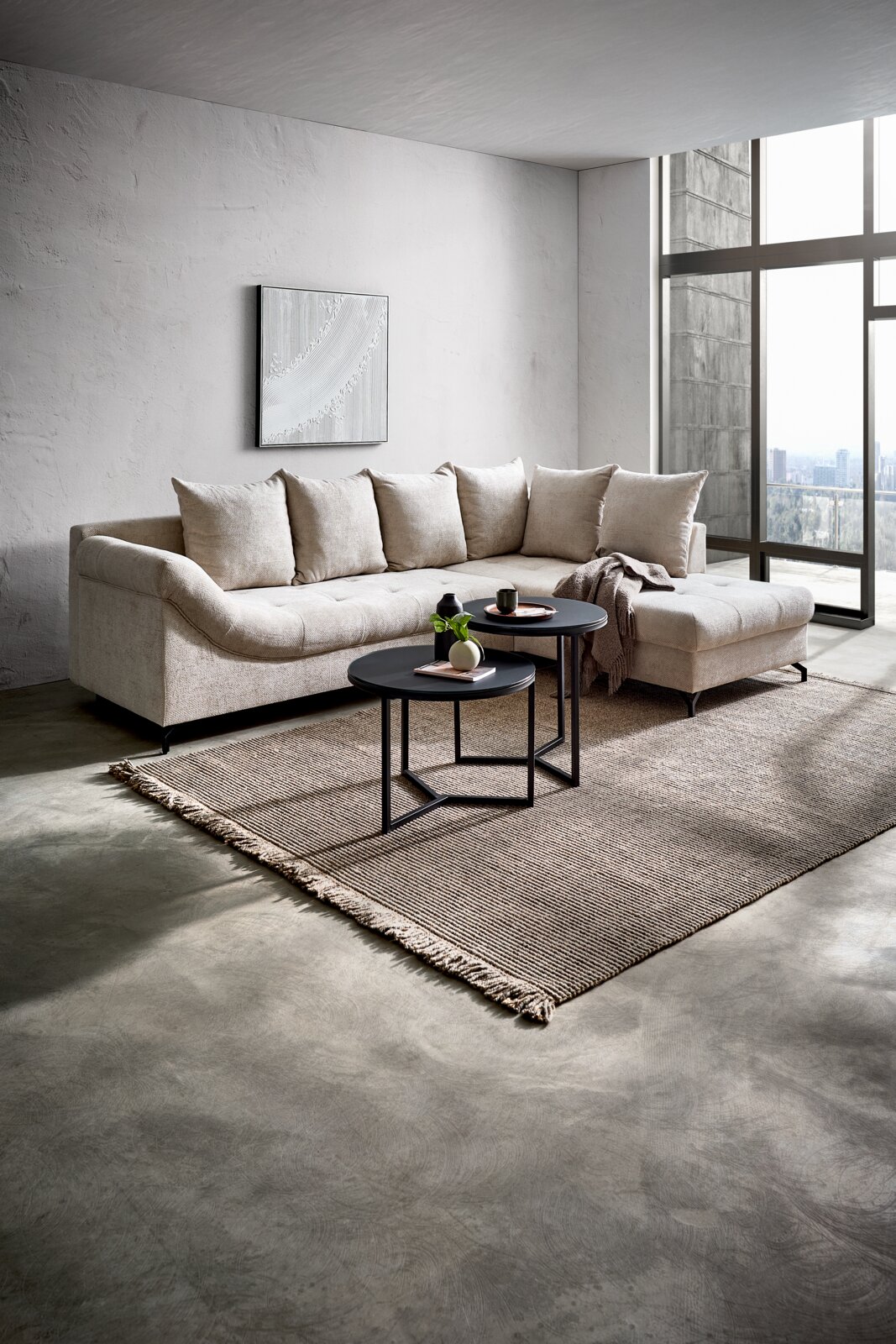 Ecksofa METZ in einem modernen Wohnzimmer mit großen Fenstern, hellem Stoff und einer Ottomane rechts. Perspektive von vorne links.