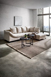 Ecksofa METZ in einem modernen Wohnzimmer mit großen Fenstern, hellem Stoff und einer Ottomane rechts. Perspektive von vorne links.