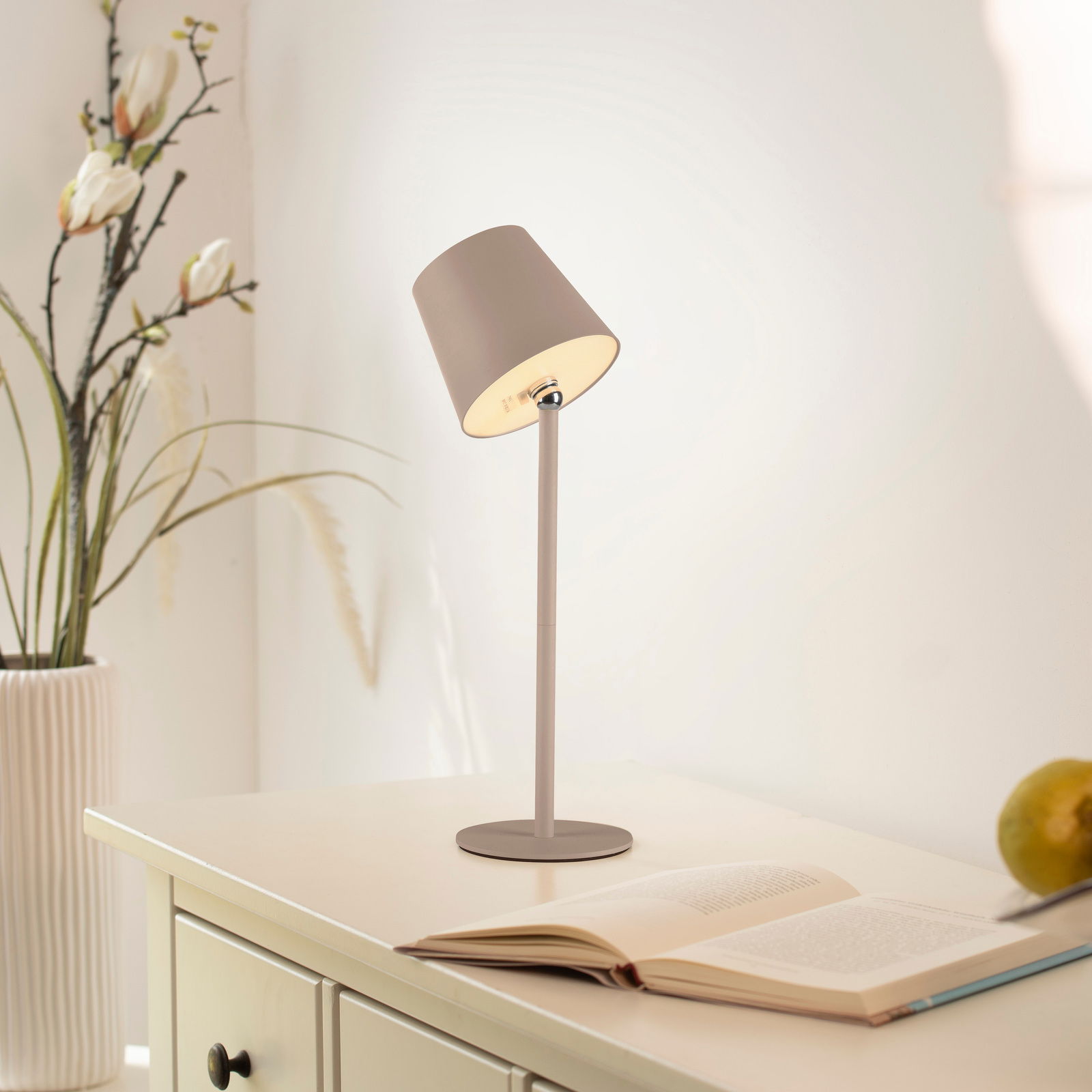Moderne Akku-Tischleuchte BOTERIA in einem hellen Raum, seitliche Perspektive, auf einem Tisch neben einem offenen Buch und einer Vase mit Blumen.