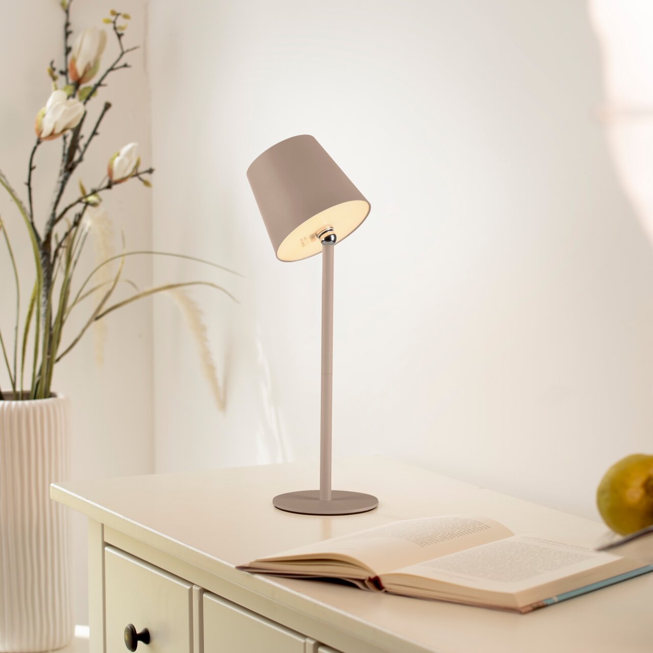 Moderne Akku-Tischleuchte BOTERIA in einem hellen Raum, seitliche Perspektive, auf einem Tisch neben einem offenen Buch und einer Vase mit Blumen.