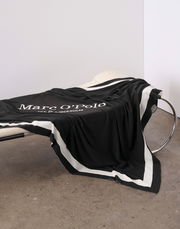 Schwarzes Plaid mit weißem Rand und Marc O'Polo Logo, drapiert auf einem modernen Stuhl, seitliche Perspektive