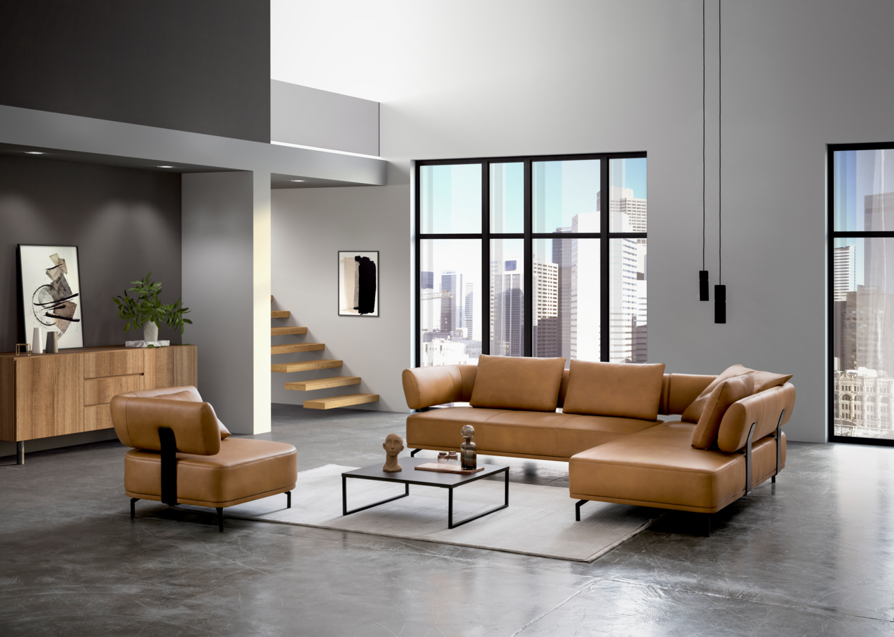 Moderne Wohnzimmermöbel in einem hellen Loft mit großen Fenstern, bestehend aus einer braunen Polsterecke und einem passenden Sessel, aus der Perspektive von vorne links betrachtet.