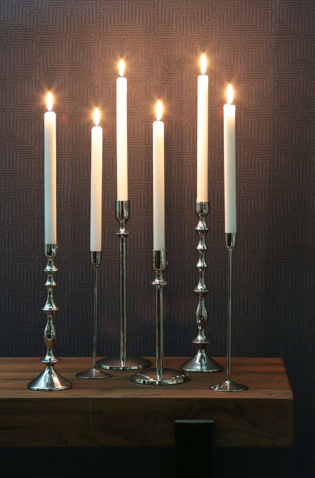 Sechs elegante silberne Kerzenständer mit brennenden weißen Kerzen, frontal auf einem Holztisch vor einer gemusterten Wand.