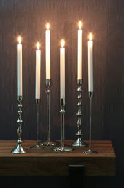 Sechs elegante silberne Kerzenständer mit brennenden weißen Kerzen, frontal auf einem Holztisch vor einer gemusterten Wand.