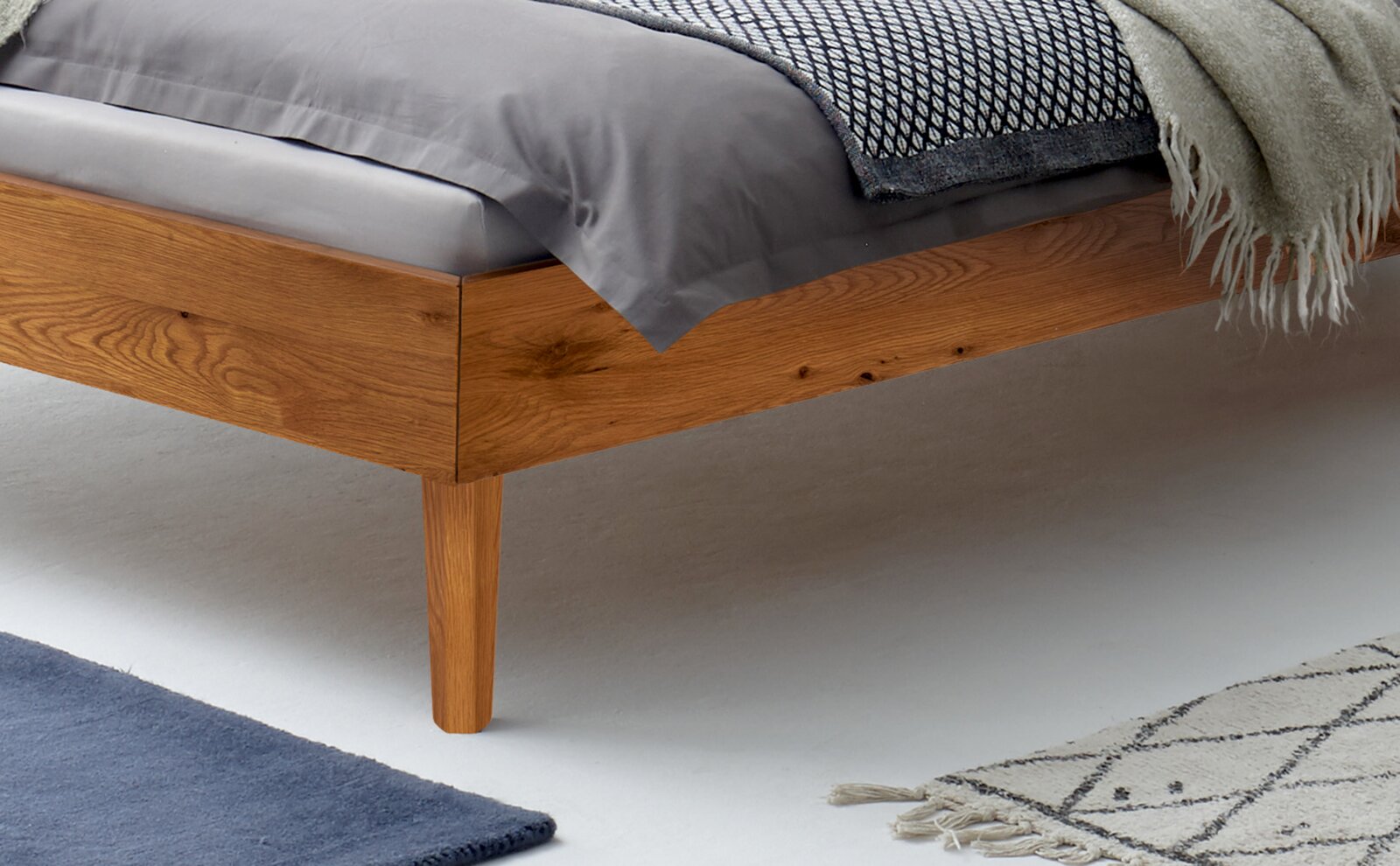 Nahaufnahme eines Bettgestells aus Holz mit Bettwäsche in Grau und einem gemusterten Teppich im Vordergrund, seitliche Perspektive