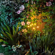 Solarbetriebene Stabbeleuchtung aus Plastik in einem Garten, umgeben von blühenden Pflanzen, seitliche Perspektive.