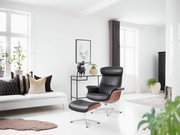 Moderne zwarte leren fauteuil met bijpassende voetenbank in een lichte woonkamer, gezien vanuit de zijkant
