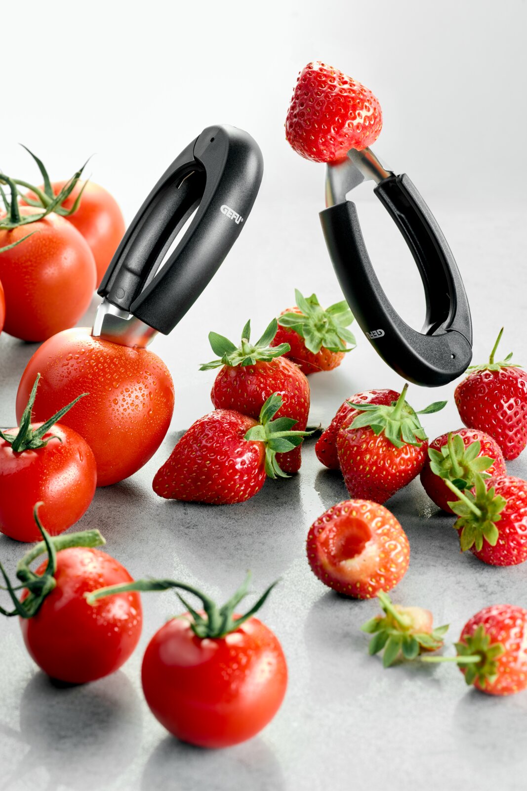 Zwarte GEFU keukenontpitter met ergonomische handgreep, schuin van voren gefotografeerd, gebruikt om een aardbei vast te klemmen; tomaten en aardbeien liggen op een lichtgrijze ondergrond.