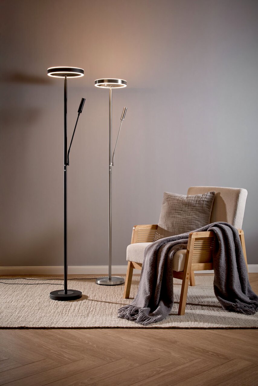 Zwei moderne LED-Deckenfluter-Stehlampen mit zusätzlichem Lesearm (schwarz und silber) neben einem Sessel im Wohnzimmer, frontale Perspektive auf Augenhöhe