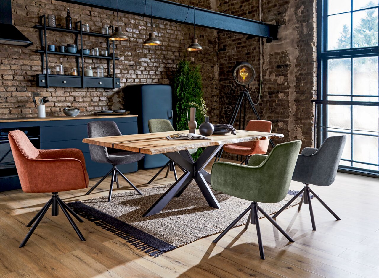 Linea Natura Esstisch 180x100 HERO Esszimmer mit modernem Esstisch aus Holz und Metallgestell, umgeben von verschiedenen farbigen Sesseln in einem industriellen Loft-Ambiente, seitliche Perspektive.