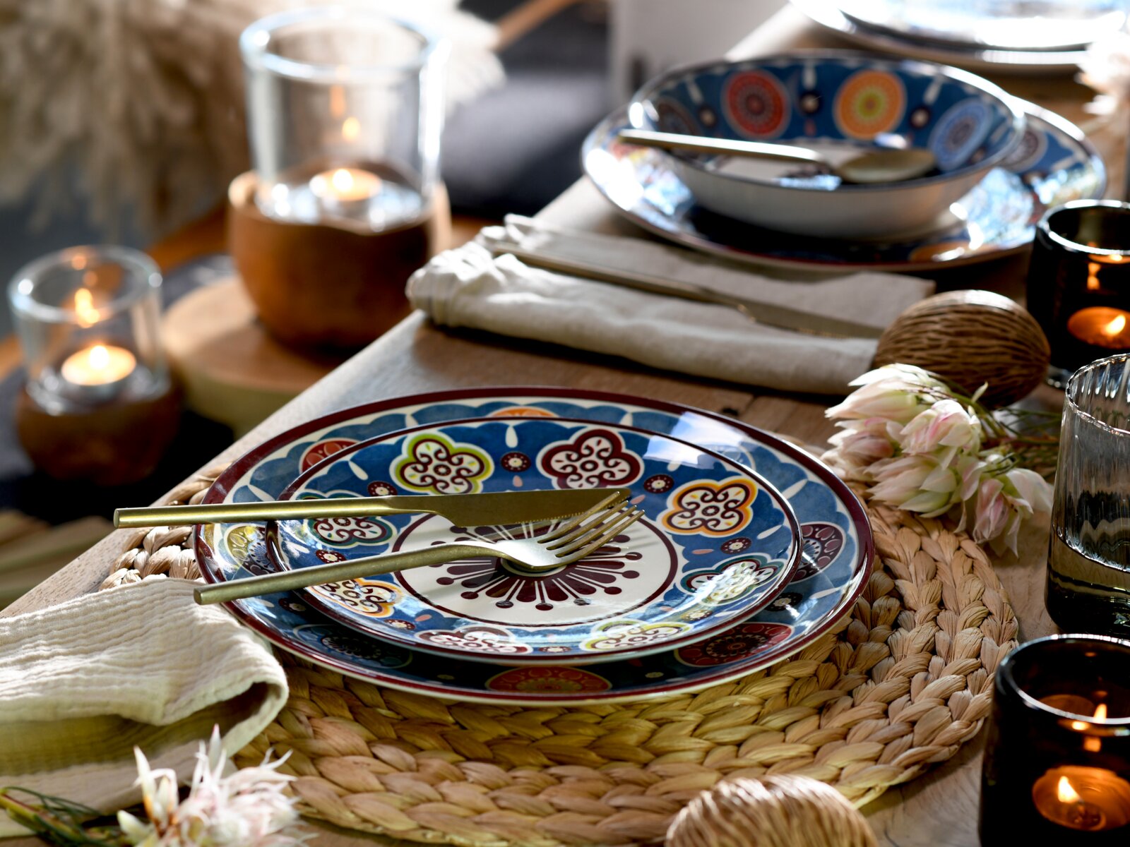 Farbenfrohes Keramik-Tafelservice mit blau-rotem Muster auf gedecktem Holztisch mit goldfarbenem Besteck, Stoffserviette, geflochtenem Platzset und Kerzenlicht, aufgenommen aus schräger Seitenansicht/leicht von oben.