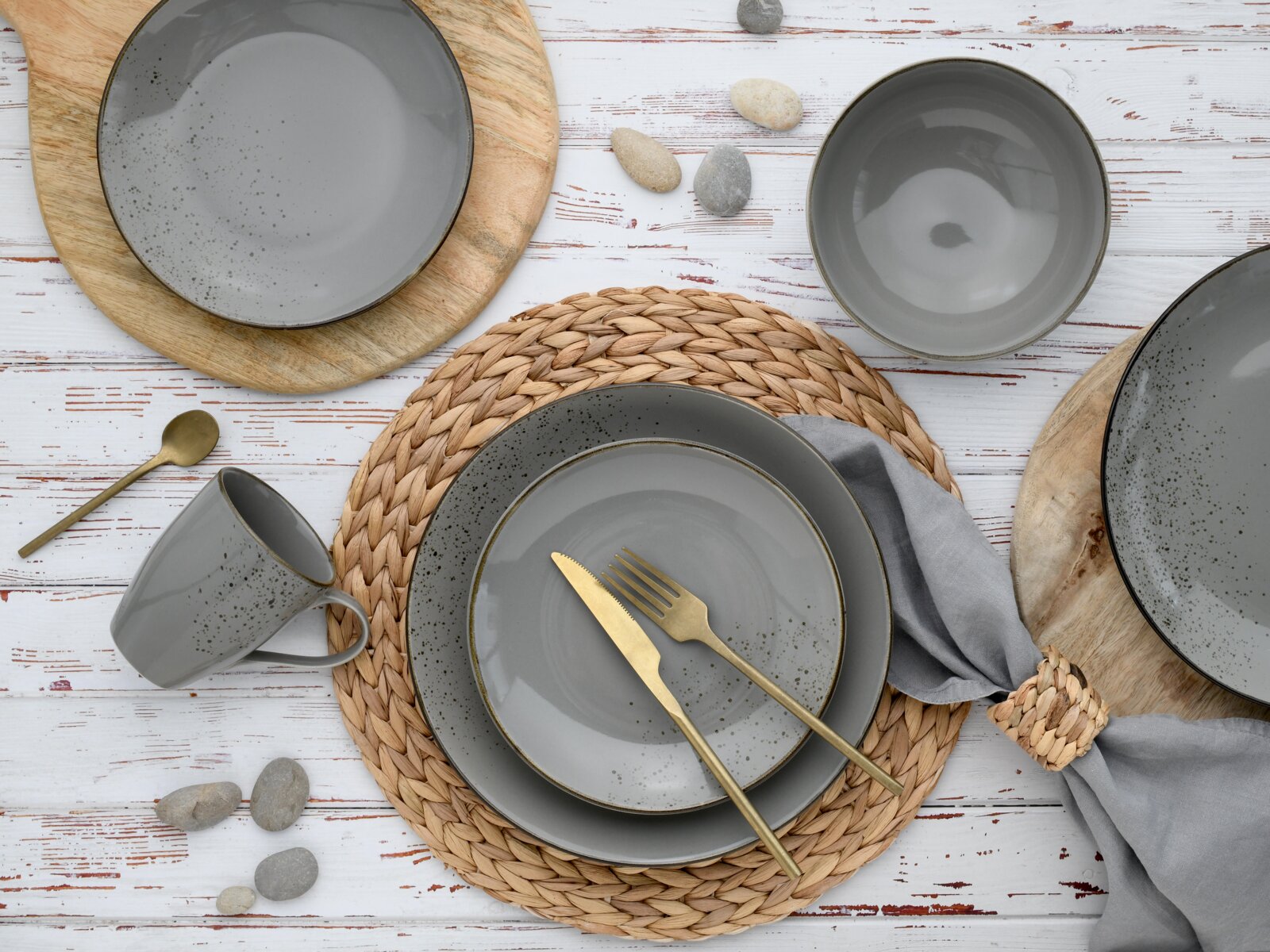 Draufsicht auf ein gedecktes Tisch-Arrangement mit grauem, gesprenkeltem Geschirr (Teller, Schalen und Tasse), goldfarbenem Besteck auf einem geflochtenen Platzset, Holzuntersetzern und grauer Stoffserviette auf weißem Holztisch.