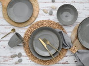 Draufsicht auf ein gedecktes Tisch-Arrangement mit grauem, gesprenkeltem Geschirr (Teller, Schalen und Tasse), goldfarbenem Besteck auf einem geflochtenen Platzset, Holzuntersetzern und grauer Stoffserviette auf weißem Holztisch.