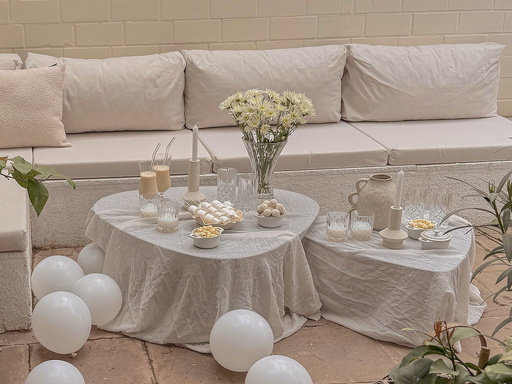 Helle Outdoor-Lounge mit weißen Sitzpolstern, dekorierten Tischen, Blumenvasen und sommerlicher Tischdeko auf einer gemütlichen Terrasse