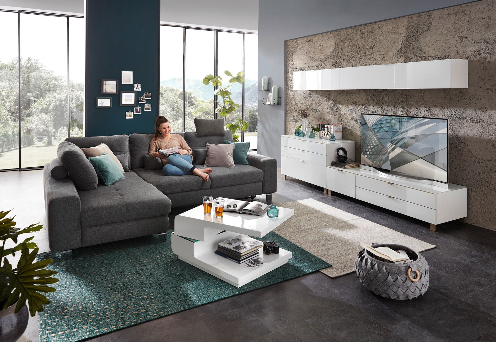 Moderne Wohnzimmermöbel in einem hellen Raum mit großen Fenstern, bestehend aus einem grauen Ecksofa, einem weißen Funktions-Couchtisch und einem weißen TV-Lowboard. Perspektive von vorne.