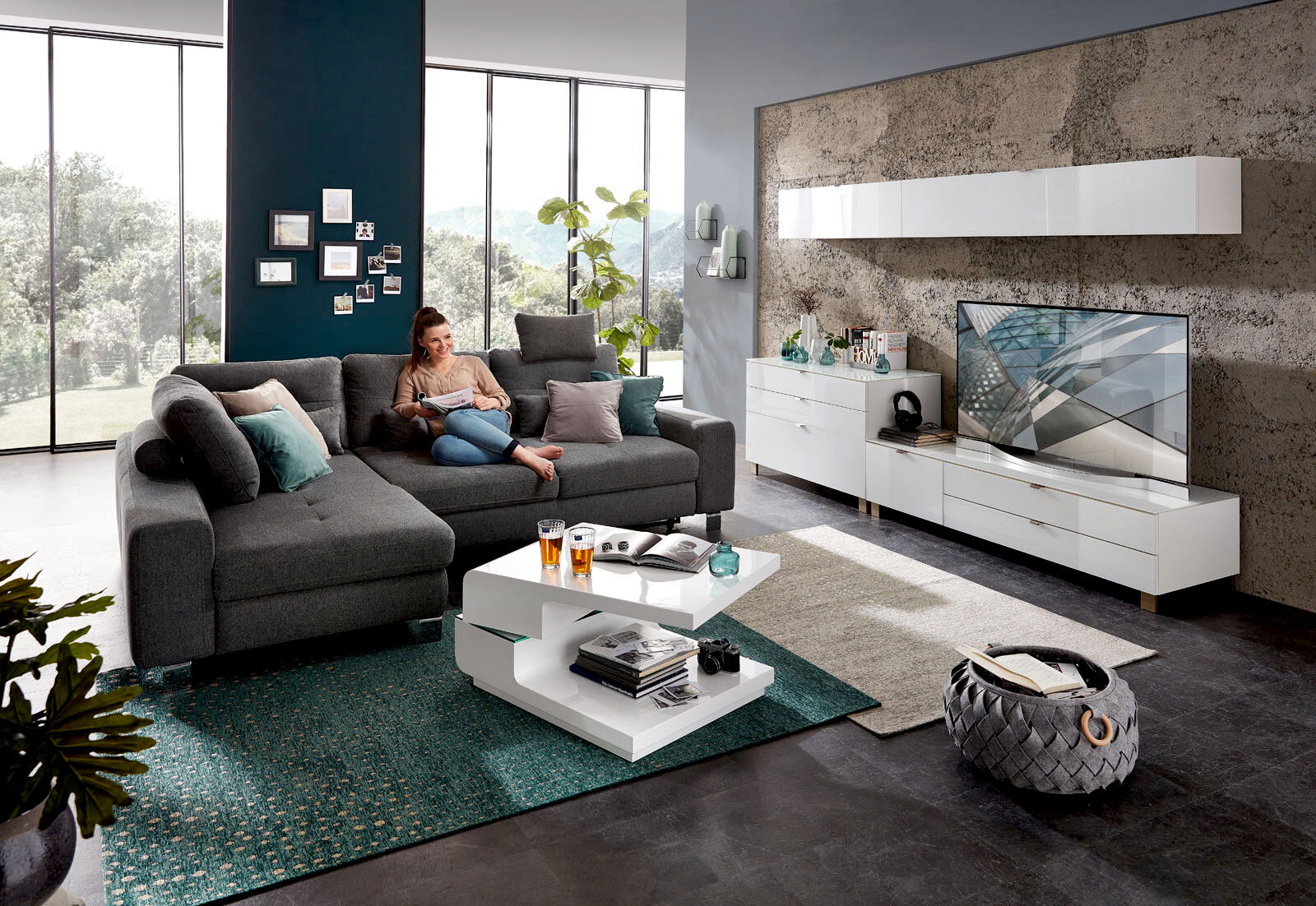 Moderne Wohnzimmermöbel in einem hellen Raum mit großen Fenstern, bestehend aus einem grauen Ecksofa, einem weißen Funktions-Couchtisch und einem weißen TV-Lowboard. Perspektive von vorne.