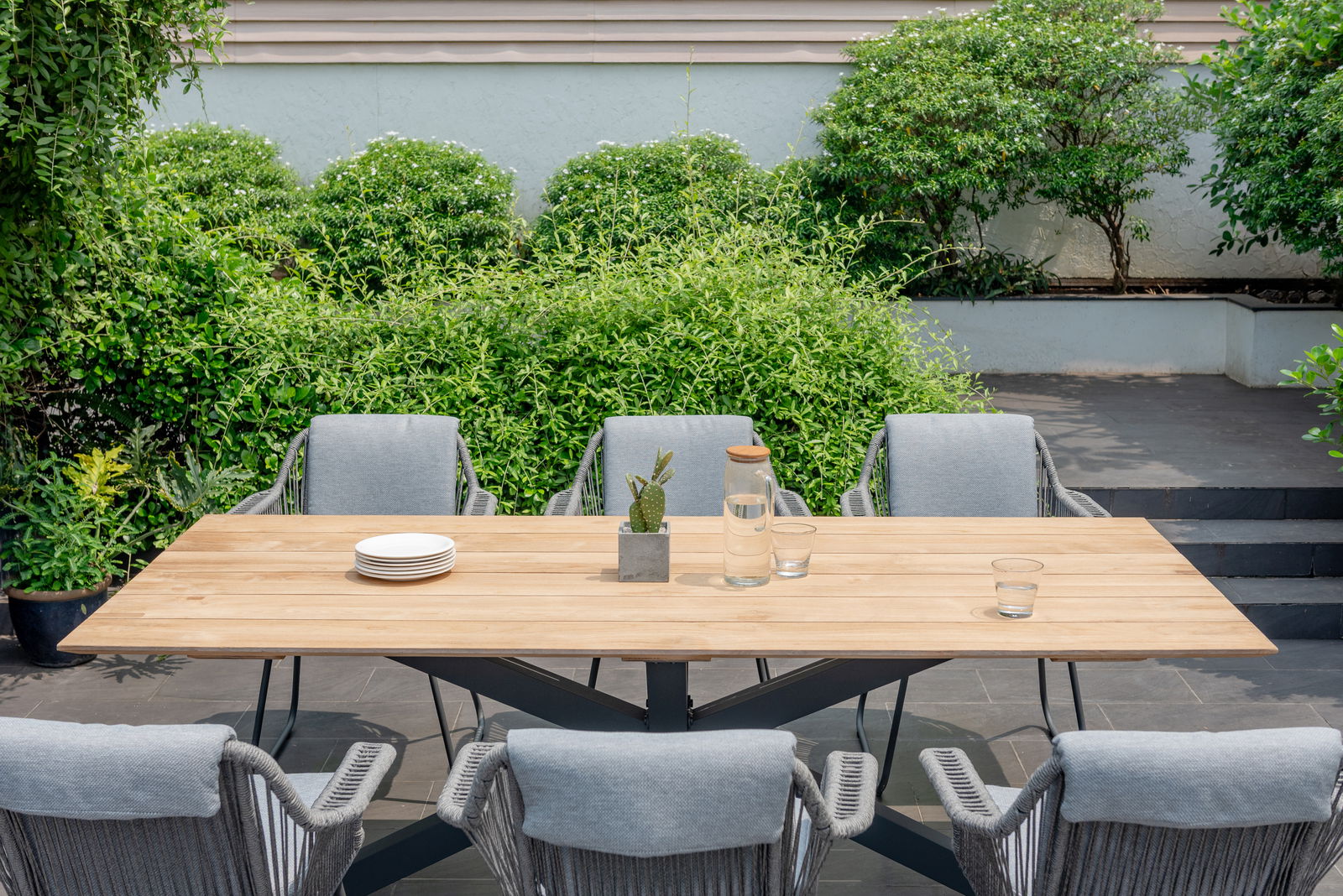 Moderner Gartentisch aus Holz mit sechs grauen Stühlen, umgeben von grünen Pflanzen, aus der Vogelperspektive fotografiert.