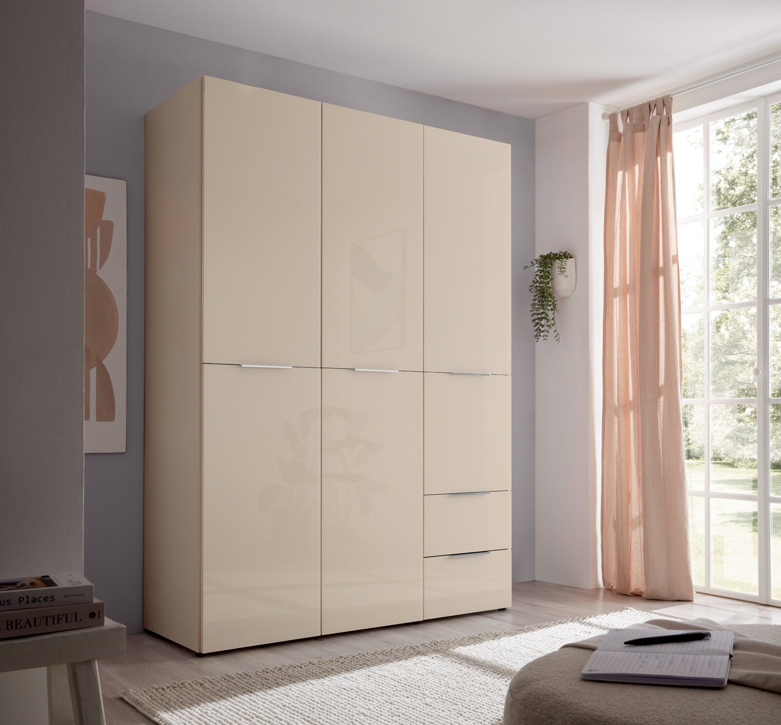 Moderner, hoher Kleiderschrank in Creme/Beige mit glatten Fronten und schmalen Griffleisten, in einem hellen Schlafzimmer neben großem Fenster mit Vorhang, perspektivisch leicht von links vorn aufgenommen.