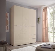 Moderner, hoher Kleiderschrank in Creme/Beige mit glatten Fronten und schmalen Griffleisten, in einem hellen Schlafzimmer neben großem Fenster mit Vorhang, perspektivisch leicht von links vorn aufgenommen.