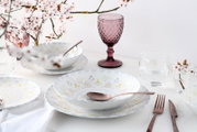 Frühlingshaft gedeckter Tisch mit weißem Geschirrset (tiefer Teller/Schale und Teller) mit pastellfarbenem Blumen- und Sternmuster, roségoldfarbenem Besteck und lila Weinglas, fotografiert in leichter Schrägansicht von oben.