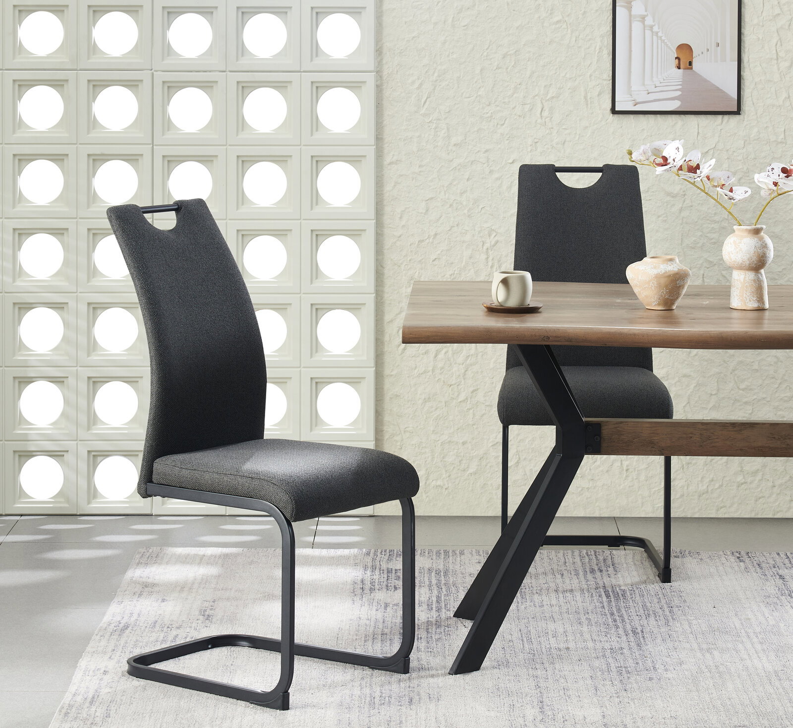 Moderner Esszimmerstuhl in Dunkelgrau mit gepolsterter Sitz- und Rückenfläche und schwarzem Freischwinger-Metallgestell, im Vordergrund schräg von vorn links fotografiert, neben Holzesstisch in hellem Wohnambiente.