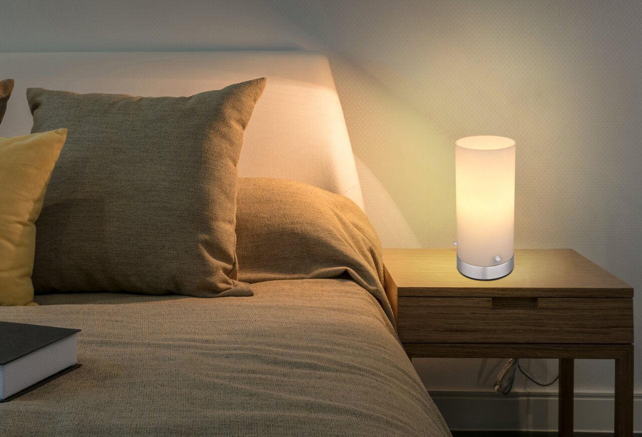 Zylindrische Tischleuchte CYLI auf einem Nachttisch neben einem Bett, seitliche Perspektive