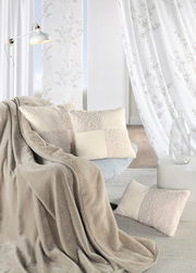 Gemütliche Wohnzimmer-Szene mit einer beigen Chenille Wohndecke, die über einem Sessel drapiert ist, und dekorativen Kissen in einem hellen, eleganten Raum mit weißen Vorhängen. Perspektive: seitliche Ansicht.