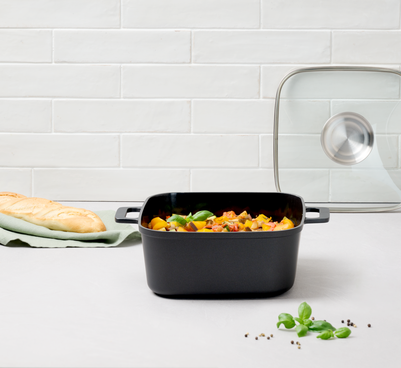 Homeware Alugussbräter quardratisch Quadratischer Alugussbräter 24cm mit Griffen, gefüllt mit Gemüse, auf einer Küchenarbeitsplatte. Im Hintergrund ein Glasdeckel und ein Baguette. Perspektive von vorne.