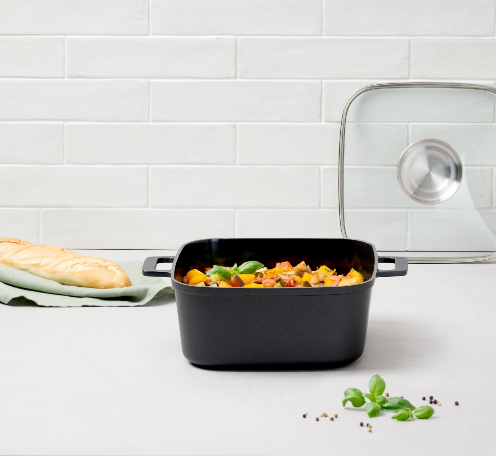 Vierkante braadpan van gegoten aluminium van 24 cm met handvatten, gevuld met groenten, op een aanrechtblad. Op de achtergrond een glazen deksel en een stokbrood. Vooraanzicht.