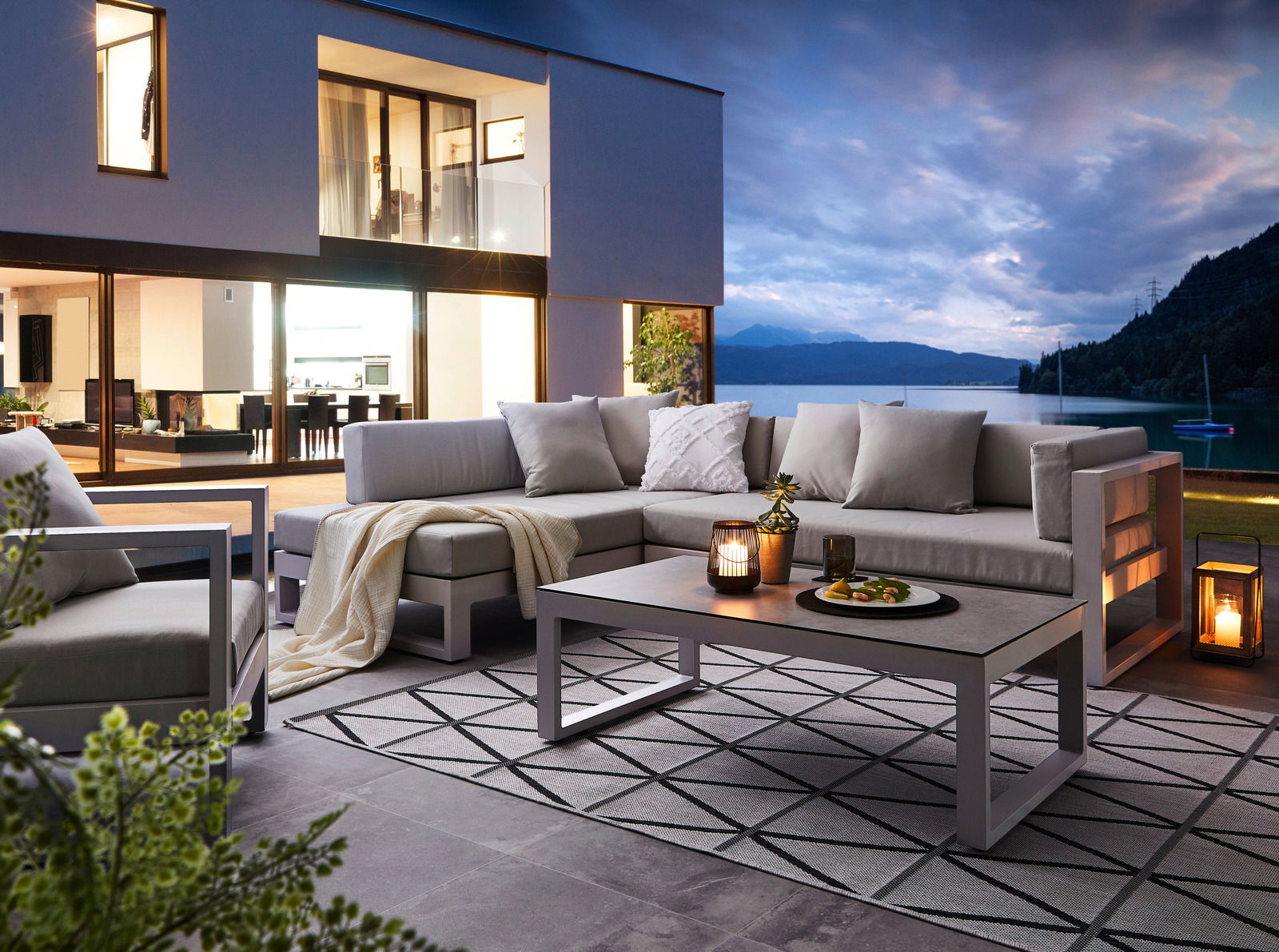 Modernes Lounge-Set auf einer Terrasse mit Blick auf einen See bei Sonnenuntergang, seitliche Perspektive.