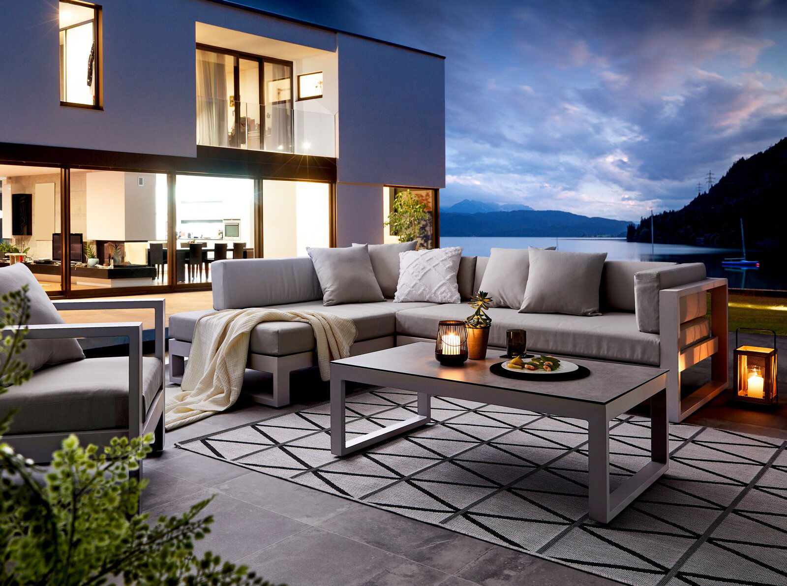 Modernes Lounge-Set auf einer Terrasse mit Blick auf einen See bei Sonnenuntergang, seitliche Perspektive.