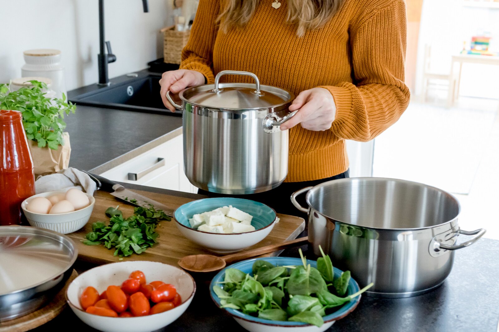 Edelstahl-Kochtopf mit Deckel und Seitengriffen wird in einer hellen Küche über der Arbeitsplatte gehalten, Vorderansicht auf den Topf; im Vordergrund Zutaten wie Tomaten, Spinat, Eier und Kräuter neben weiteren Töpfen.