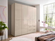 Beiger Kleiderschrank mit hochglänzenden Fronten, vier Türen und drei Schubladen im hellen Schlafzimmer neben großem Fenster, Ansicht schräg von vorne links.