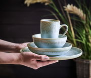 Ein stilvolles, 20-teiliges Kombiservice in sanften Grüntönen, bestehend aus Tasse, Schale und Teller, wird von Händen präsentiert. Die Perspektive zeigt das Set von der Seite, mit einem natürlichen Hintergrund aus Pflanzen.