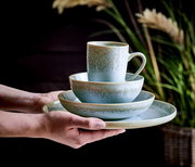 Ein stilvolles, 20-teiliges Kombiservice in sanften Grüntönen, bestehend aus Tasse, Schale und Teller, wird von Händen präsentiert. Die Perspektive zeigt das Set von der Seite, mit einem natürlichen Hintergrund aus Pflanzen.