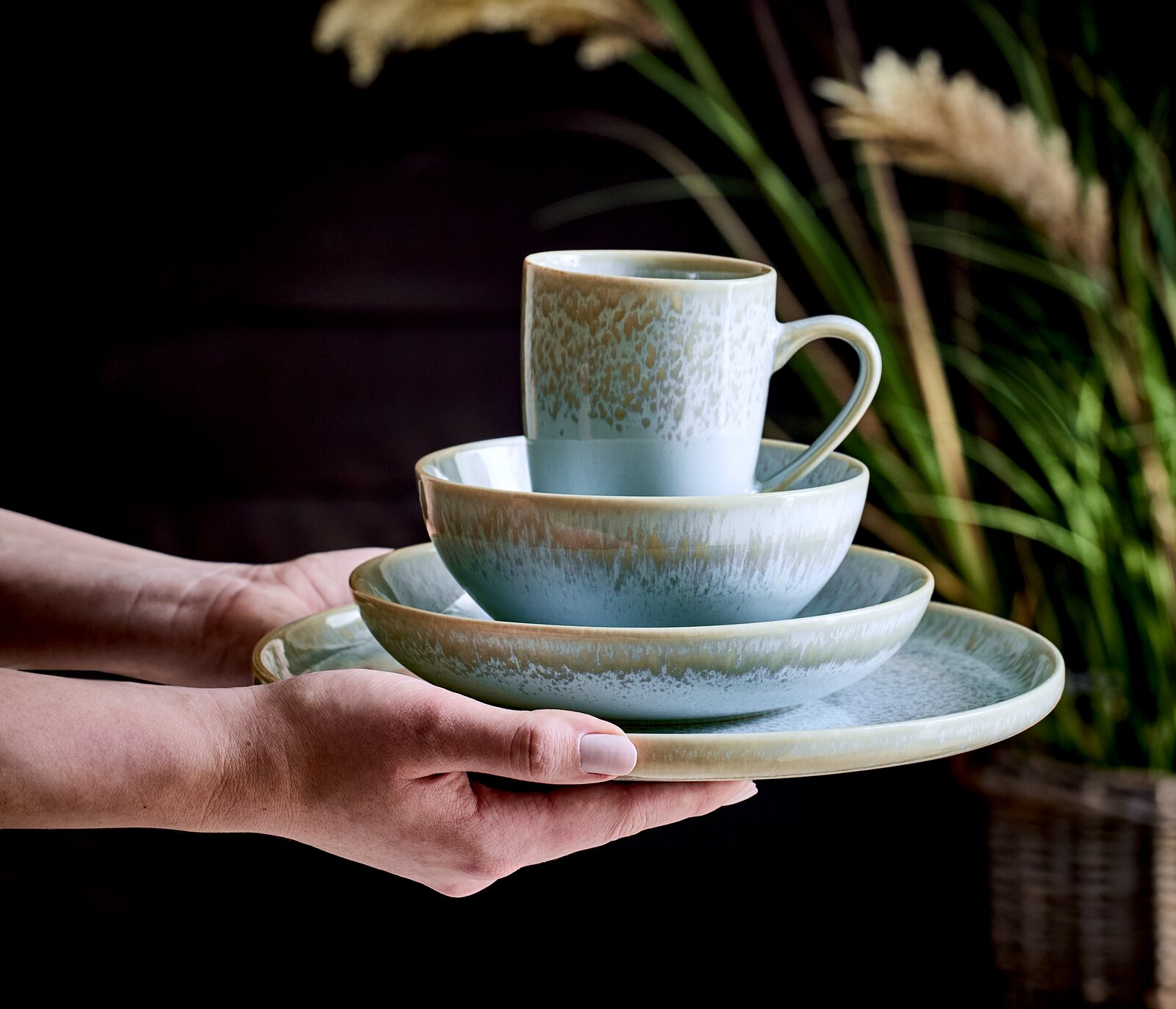 Ein stilvolles, 20-teiliges Kombiservice in sanften Grüntönen, bestehend aus Tasse, Schale und Teller, wird von Händen präsentiert. Die Perspektive zeigt das Set von der Seite, mit einem natürlichen Hintergrund aus Pflanzen.