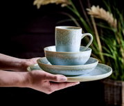 Ein stilvolles, 20-teiliges Kombiservice in sanften Grüntönen, bestehend aus Tasse, Schale und Teller, wird von Händen präsentiert. Die Perspektive zeigt das Set von der Seite, mit einem natürlichen Hintergrund aus Pflanzen.