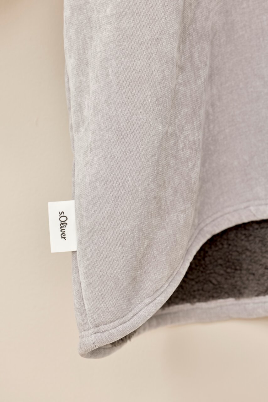 Nahaufnahme eines grauen Lounge Hoodies mit weichem Innenfutter und sichtbarem Markenetikett, seitliche Perspektive.