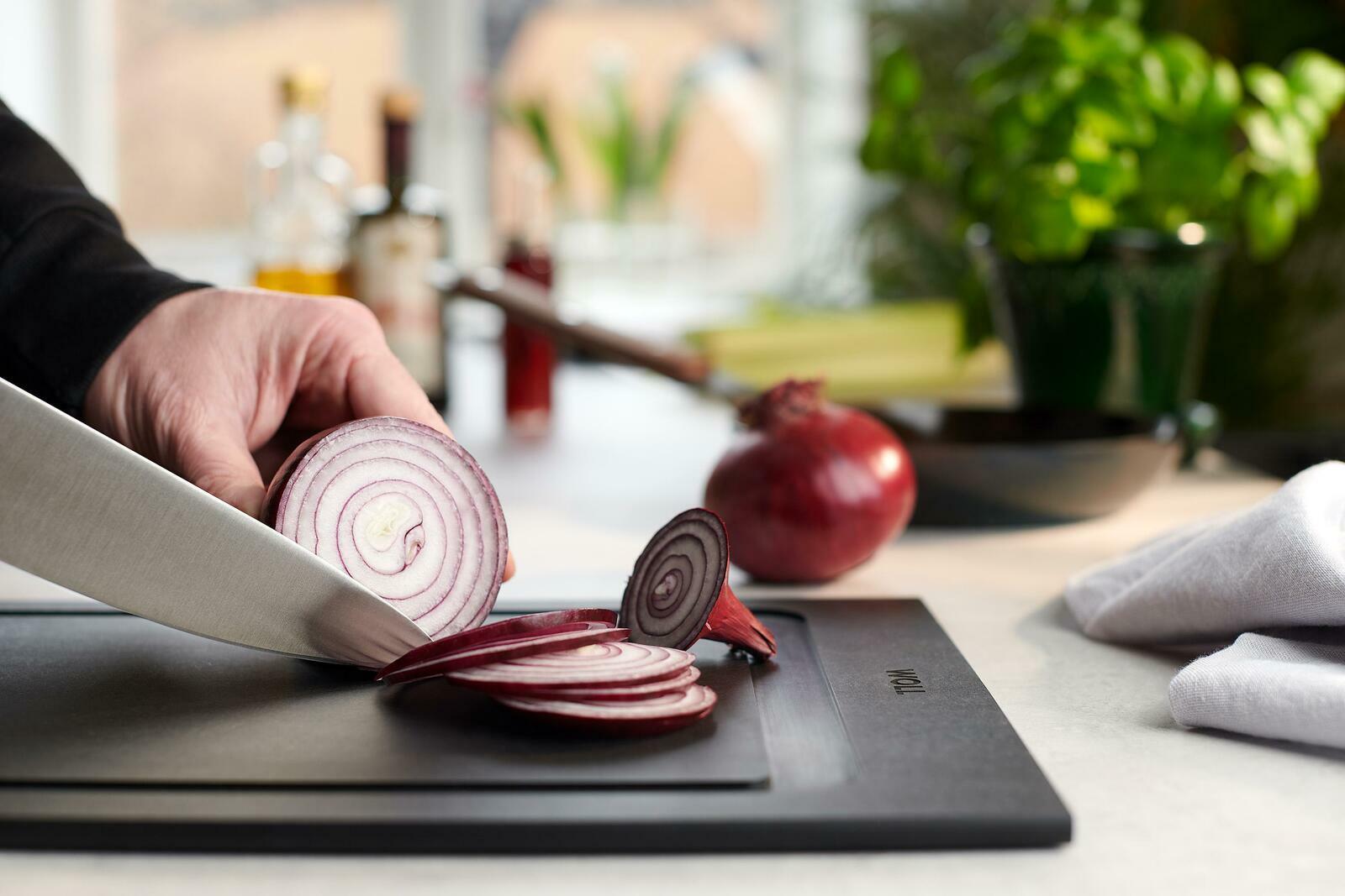 Nahaufnahme eines Schneidebretts mit einer Hand, die eine rote Zwiebel in Scheiben schneidet. Im Hintergrund sind Küchenutensilien und Pflanzen zu sehen.